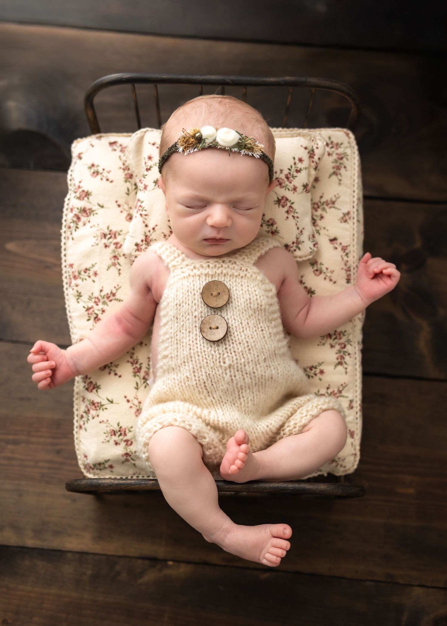 Sleeping newborn baby in cream knit romper with floral headband lying on vintage miniature bed with floral blanket.