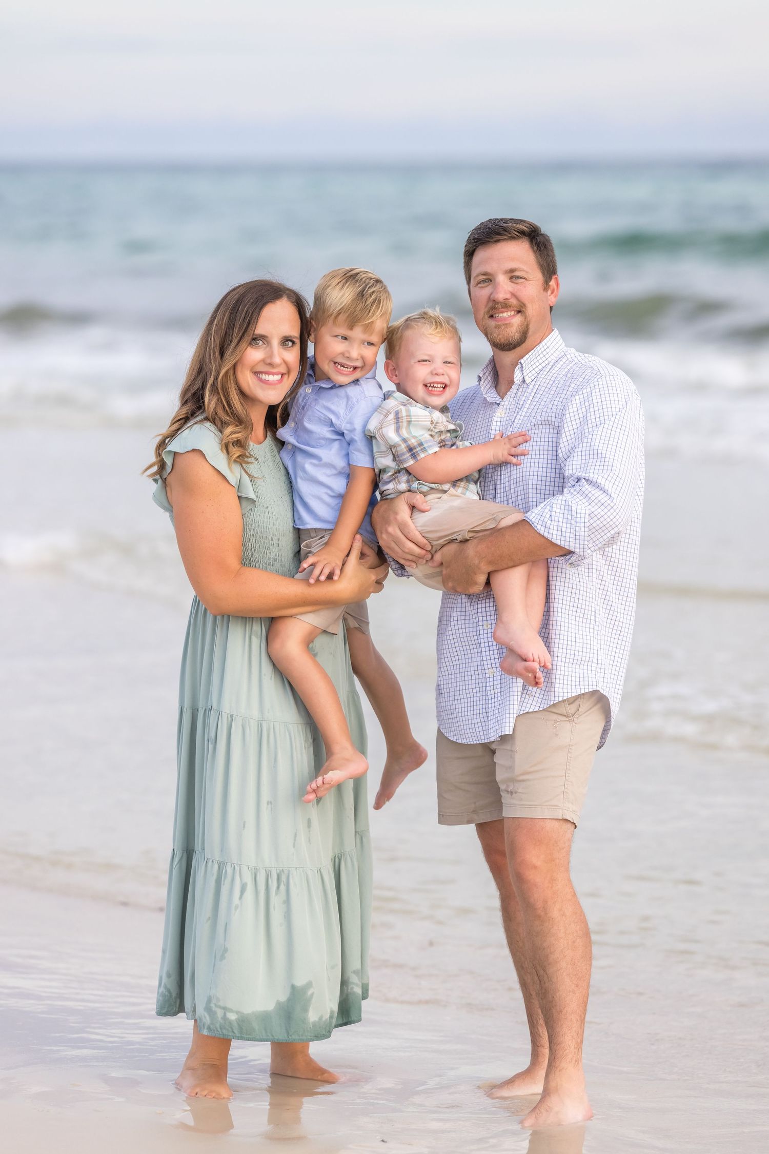 Family of four poses on a beach, parents holding two young boys, wearing coordinated outfits in soft blues and greens.