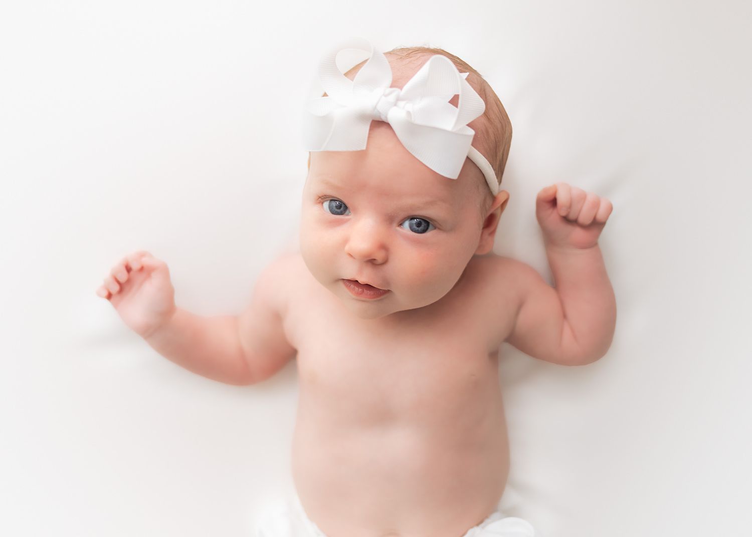 Baby wearing white bow headband lying on white background.
