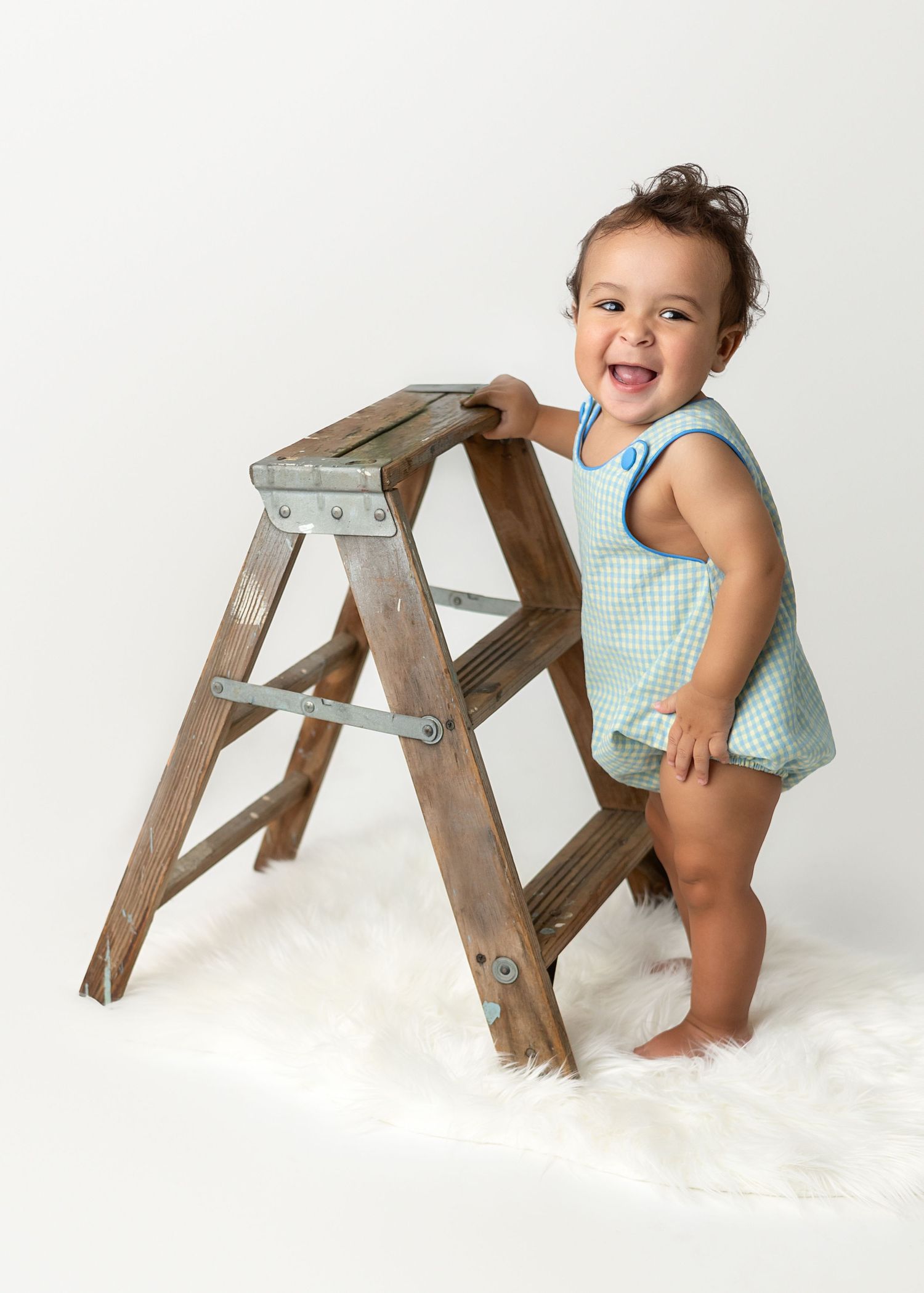 Baby in blue checkered outfit poses with wooden stepladder on white background.