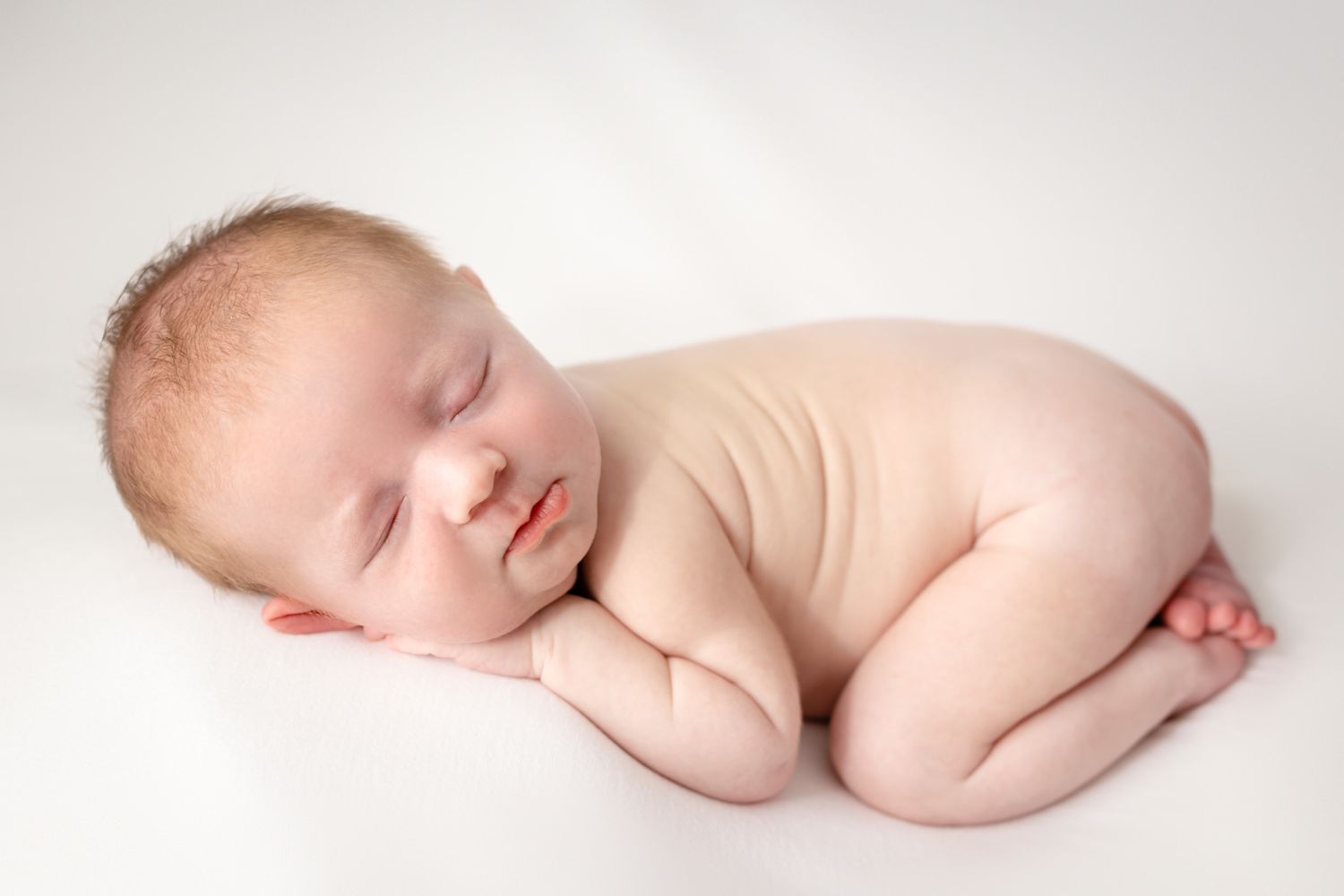 Sleeping newborn posed in fetal position on white backdrop.