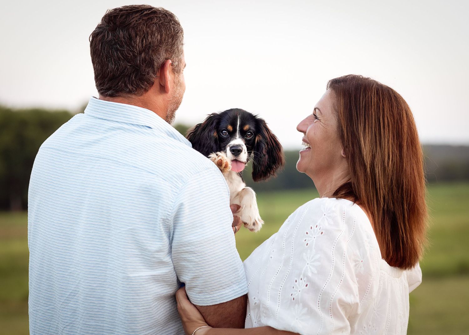 Joyful couple embraces while holding their spotted puppy in a secluded Alabama field for photo shoot.