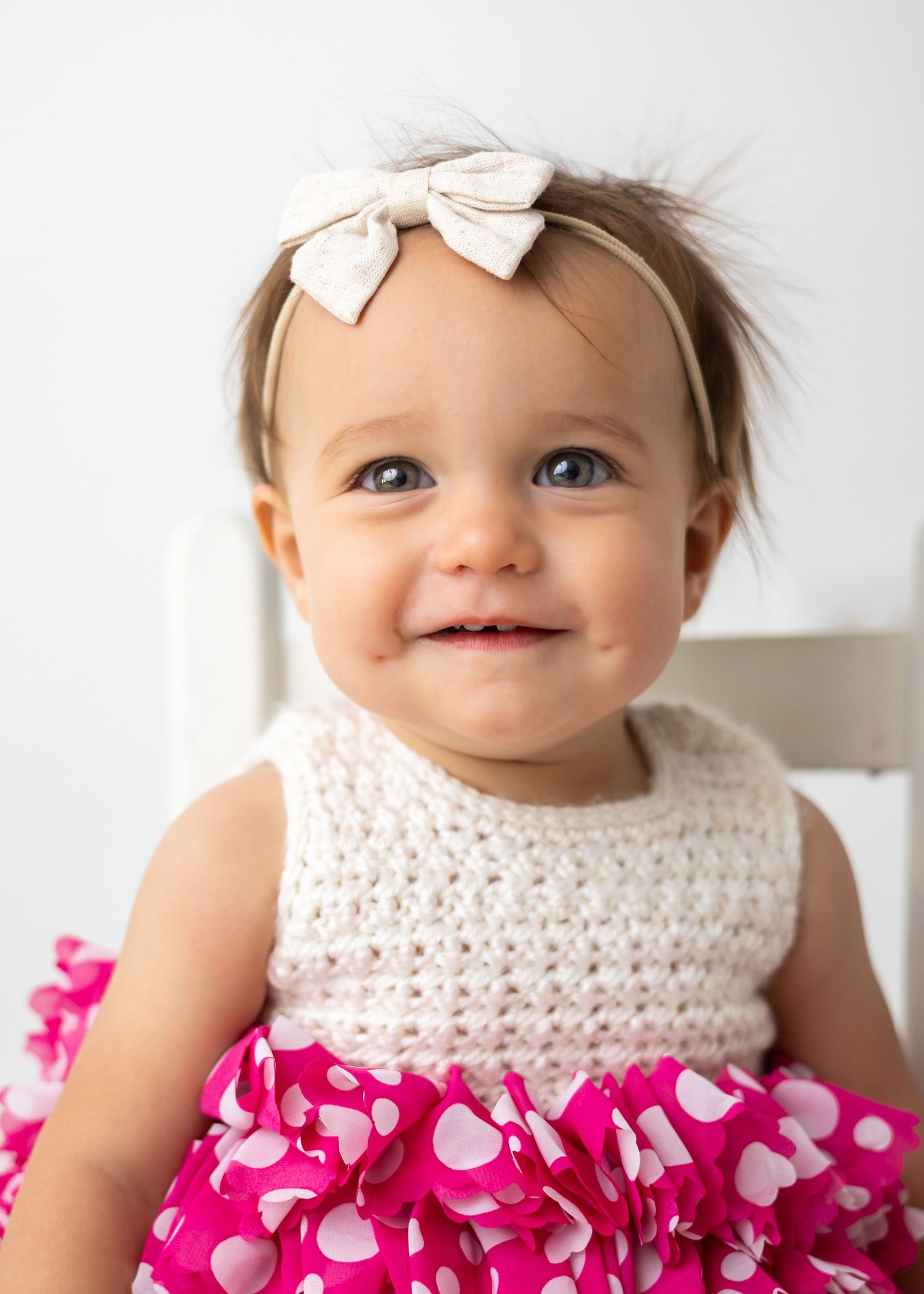 Smiling baby girl wearing a cream crochet top, pink polka dot dress, and bow headband, sitting in a white chair.