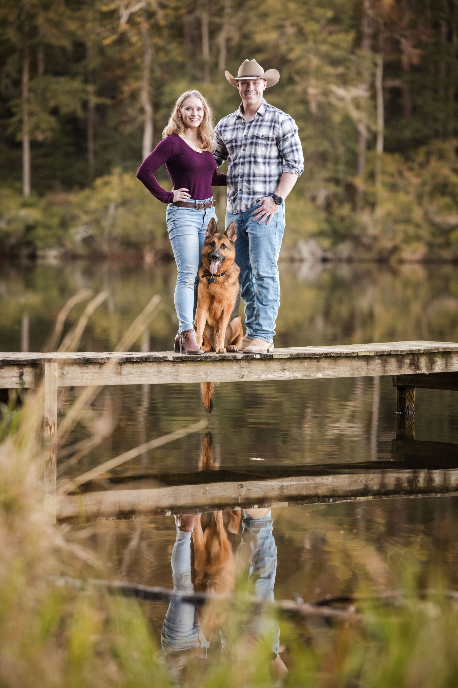 A couple and their German Shepherd dog pose on a wooden dock by a serene lake during an outdoor photo session.