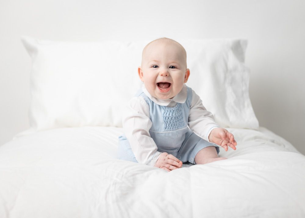 Happy baby in light blue overalls sits on white bedding while smiling at the camera.