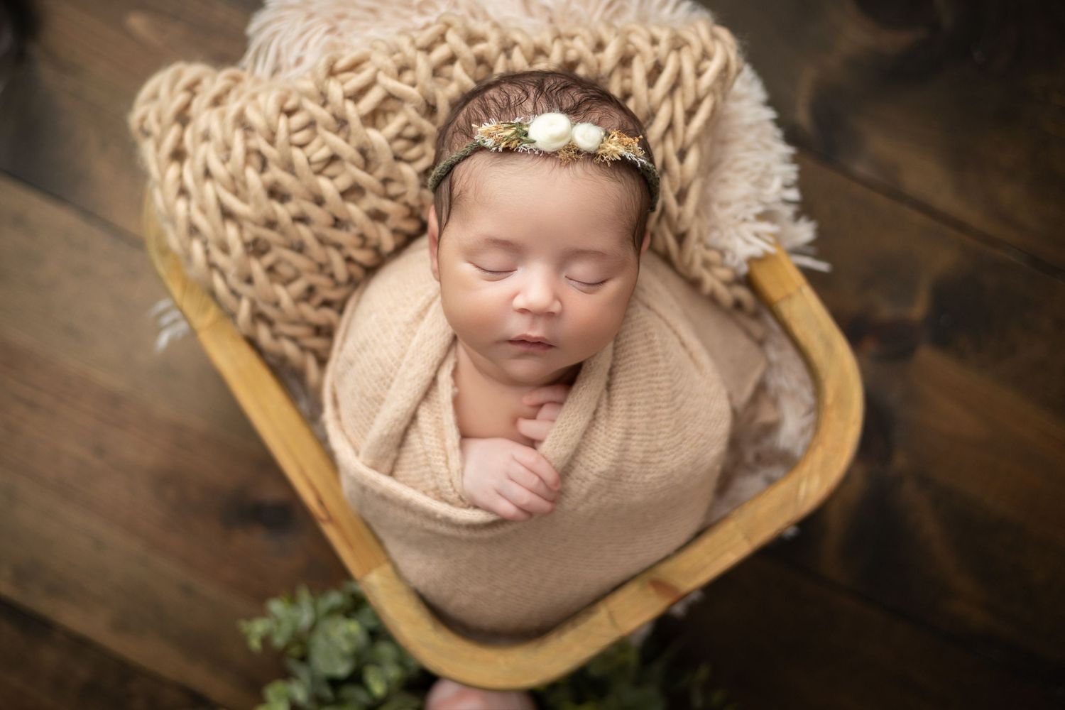 Sleeping newborn baby wrapped in beige knit blanket with floral headband in wooden basket on dark wood floor Daleville, AL studio