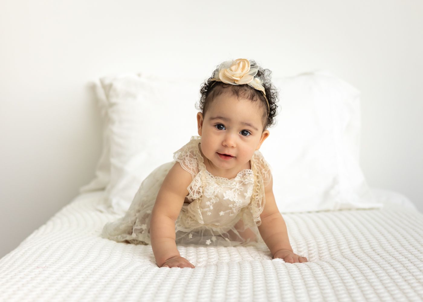 A baby in a white lace dress crawls on white bedding while looking at the camera.