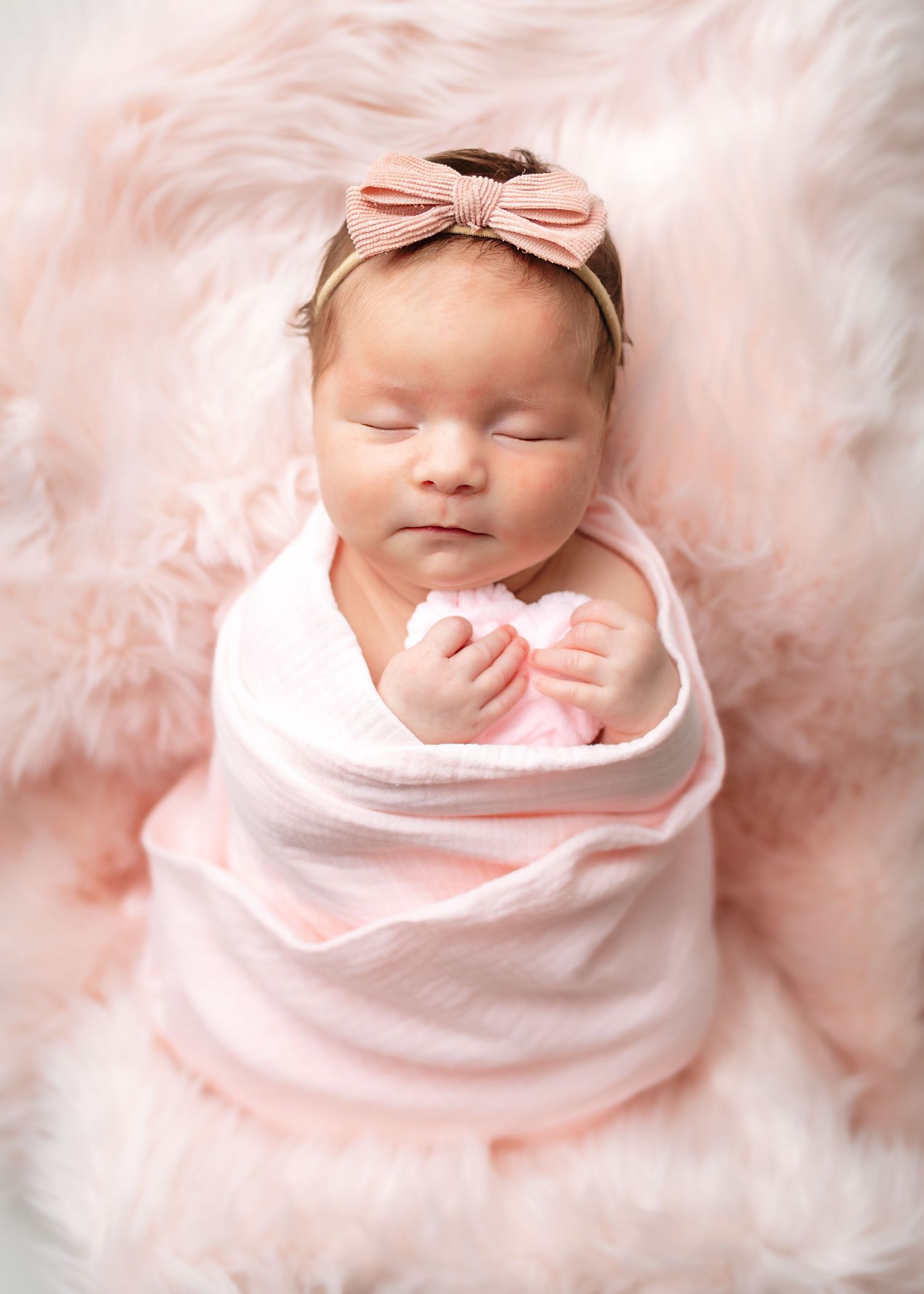 Newborn baby swaddled in pink wrap with bow headband on soft pink background.