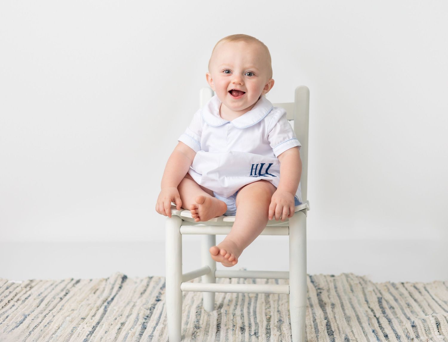 Joyful baby in white outfit sitting on small white chair during first year portraits.