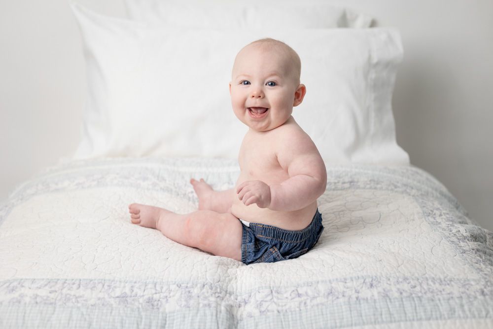 Six month old baby boy wearing navy shorts sits upright on white bed.