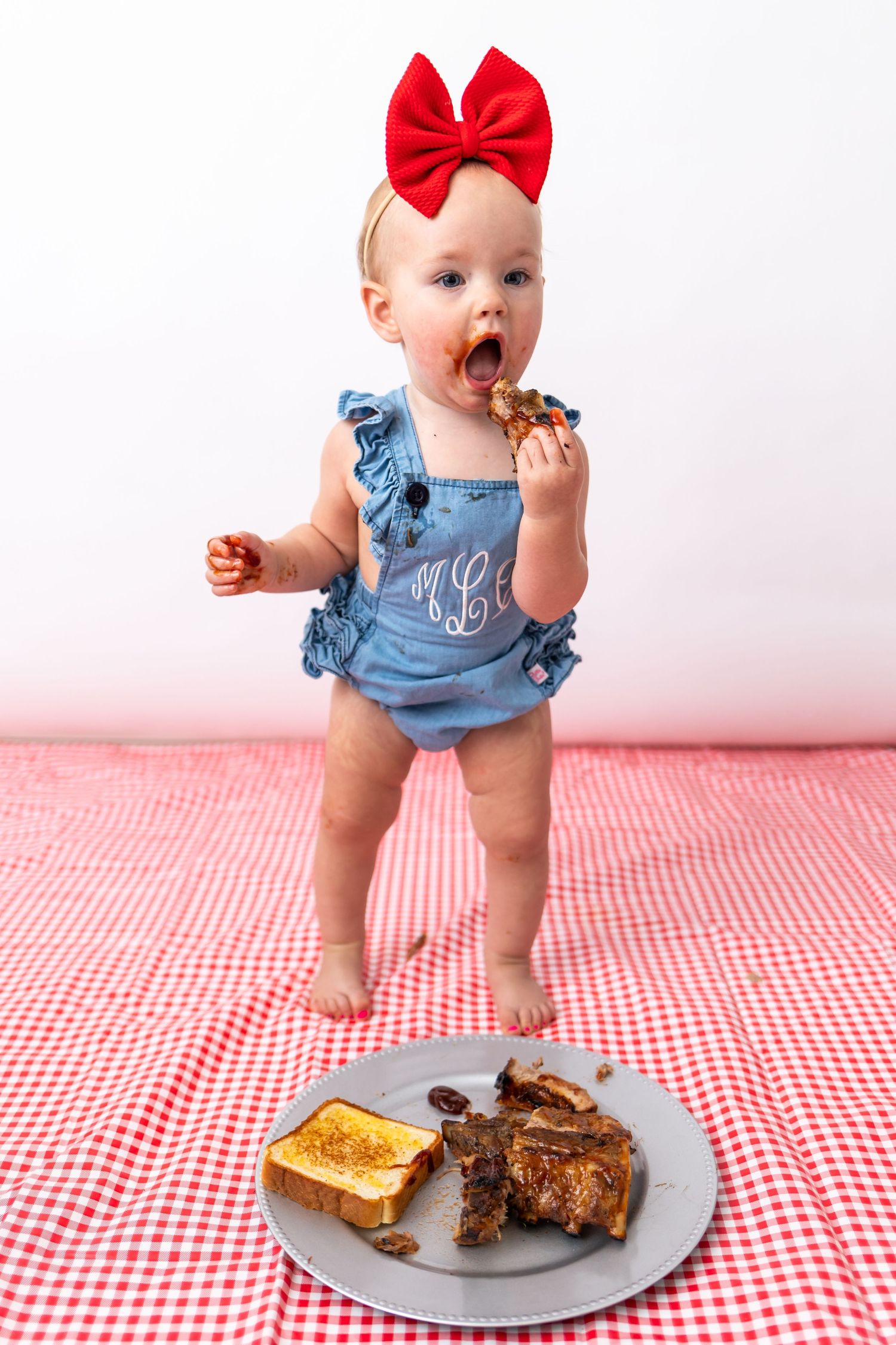 Baby in denim overalls and red bow reaching for toast and brownies on checkered tablecloth.