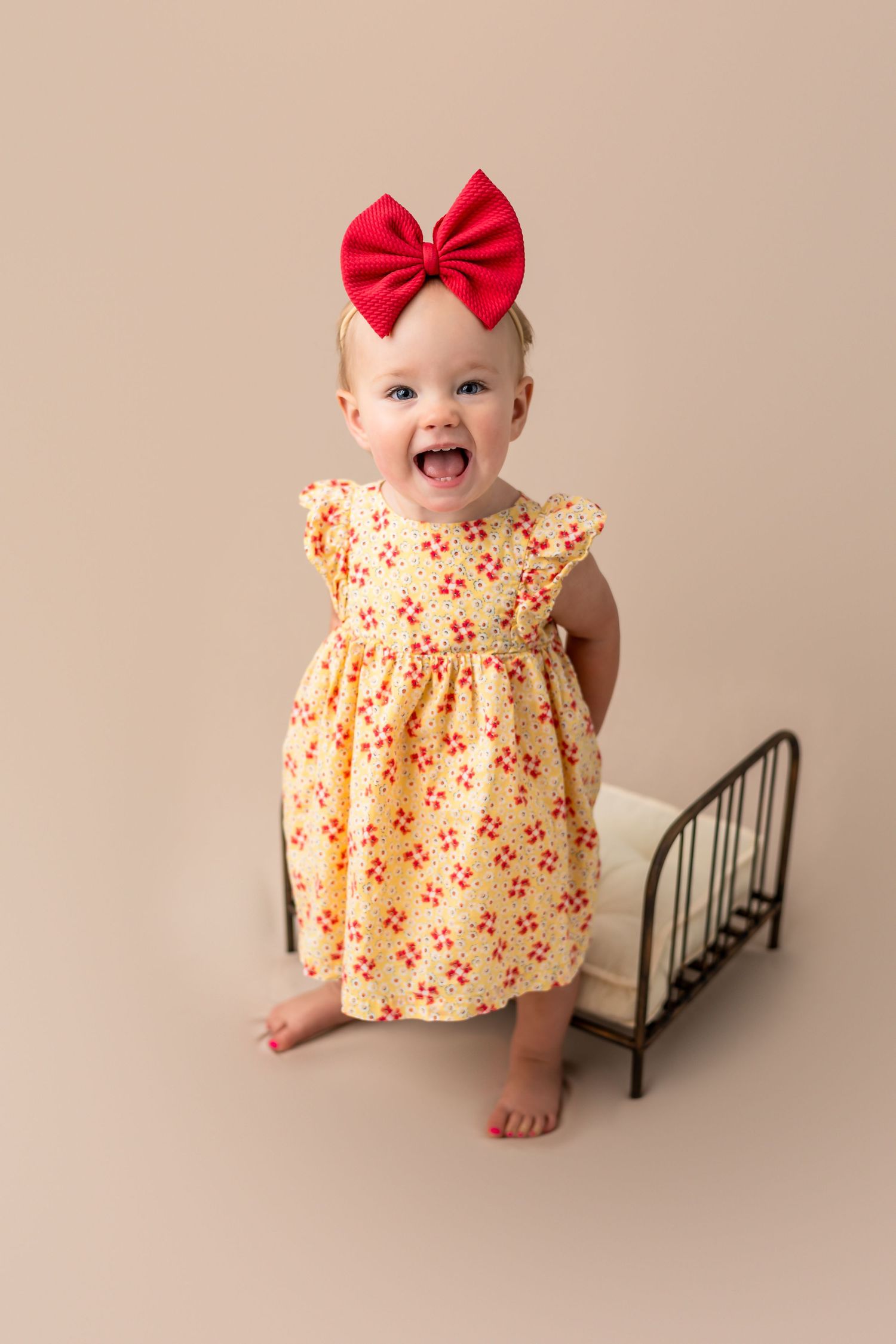 Baby girl poses in a yellow floral dress with red bow next to a small metal bed frame for photography shoot.