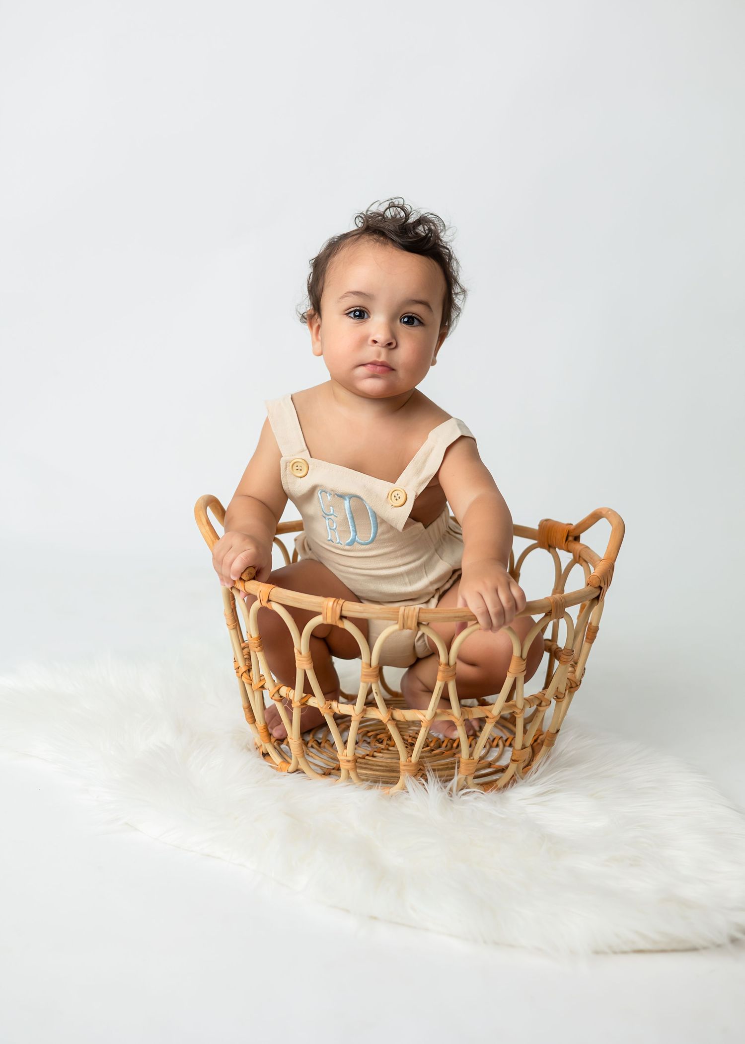 Baby sits in woven basket on white fluffy rug during photo session.