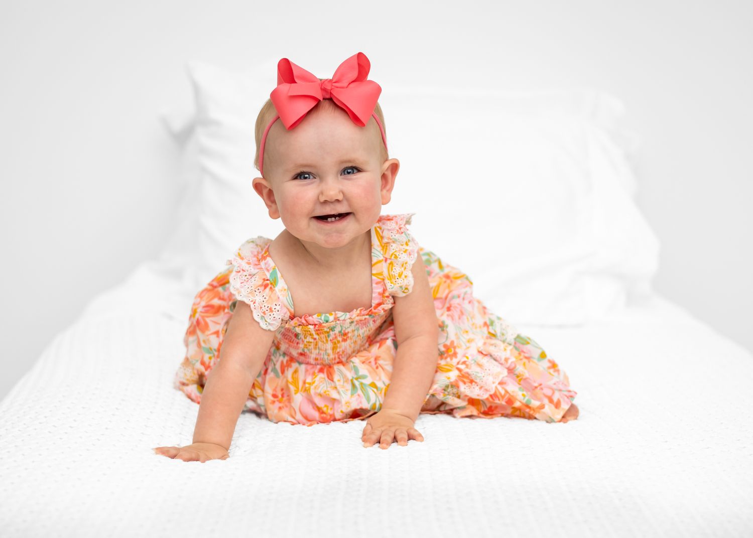 Baby in floral dress crawls and smiles on white blanket in studio setting.
