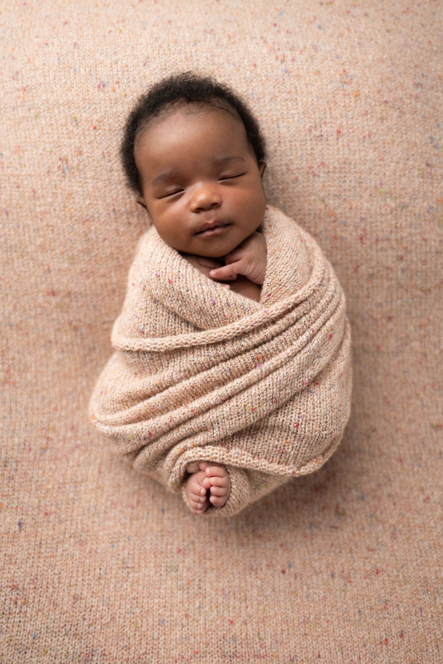 Sleeping newborn wrapped in beige knit blanket on textured background.