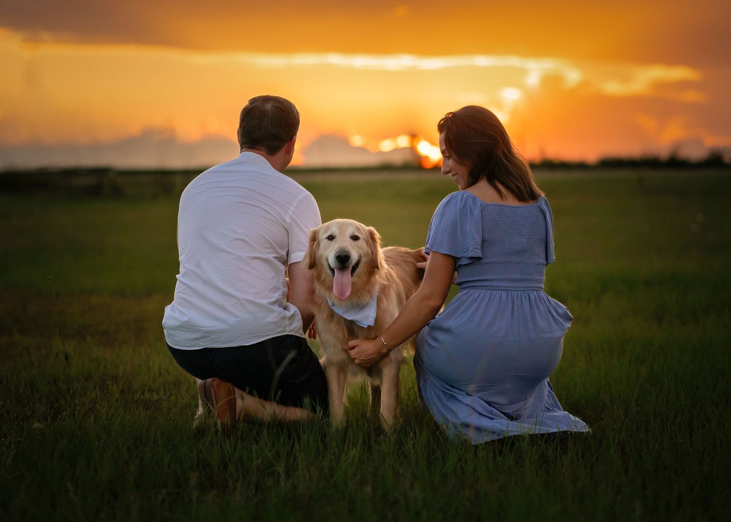 A couple and their dog sit together watching a beautiful golden sunset in a grassy field during their photo shoot.