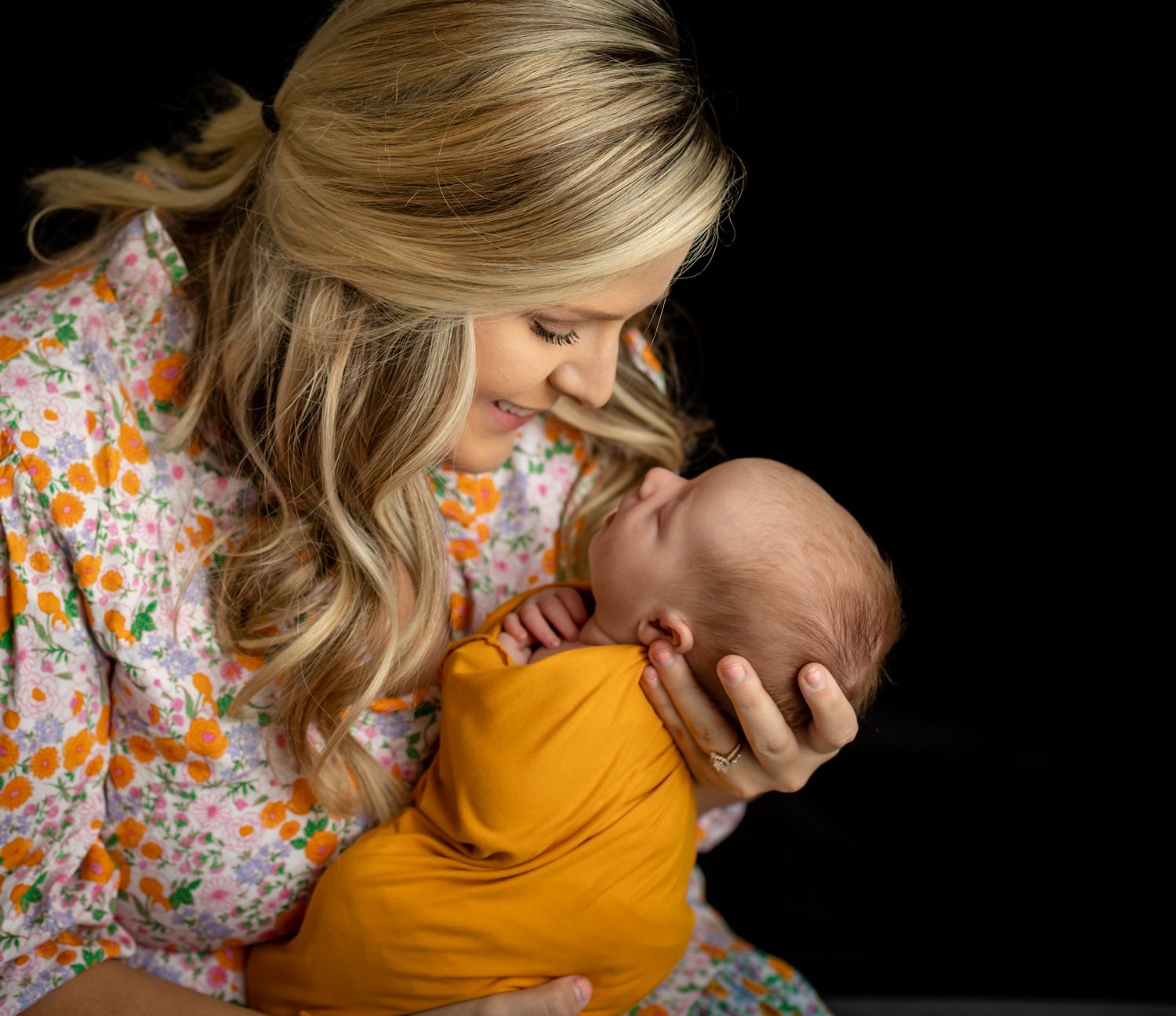 Mother in floral dress holding baby wrapped in mustard yellow swaddle.