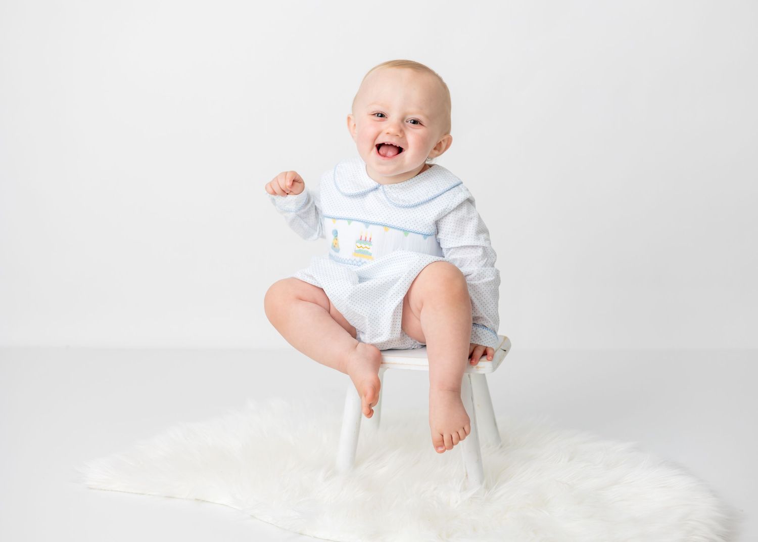 Happy baby in white outfit sitting on small white chair against light background.