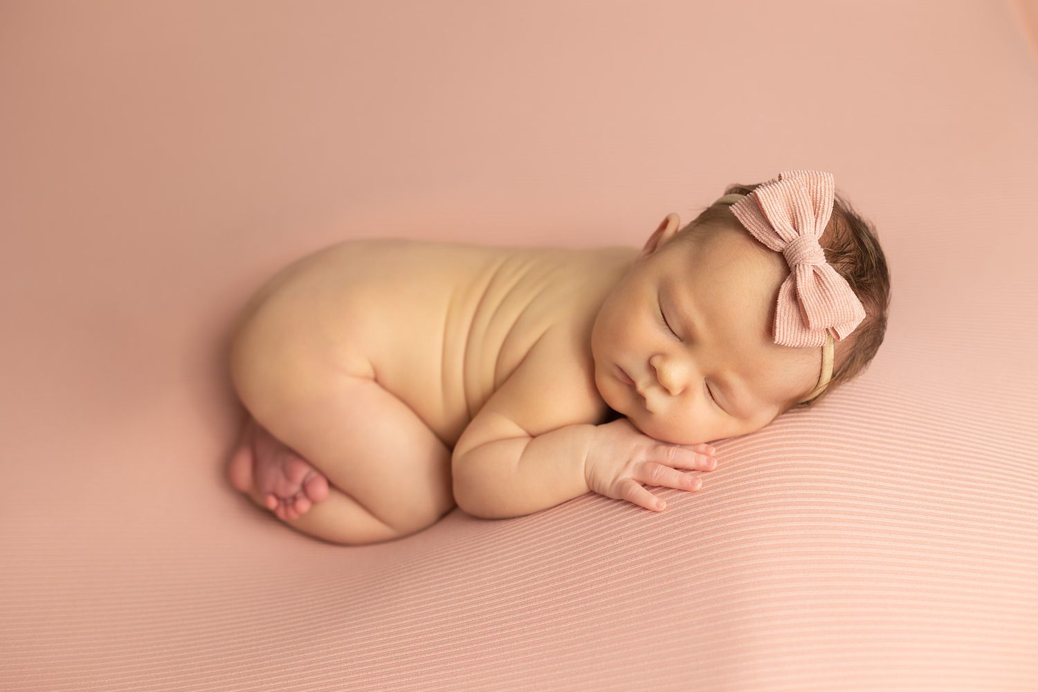 Sleeping newborn with pink bow headband poses on pink background.