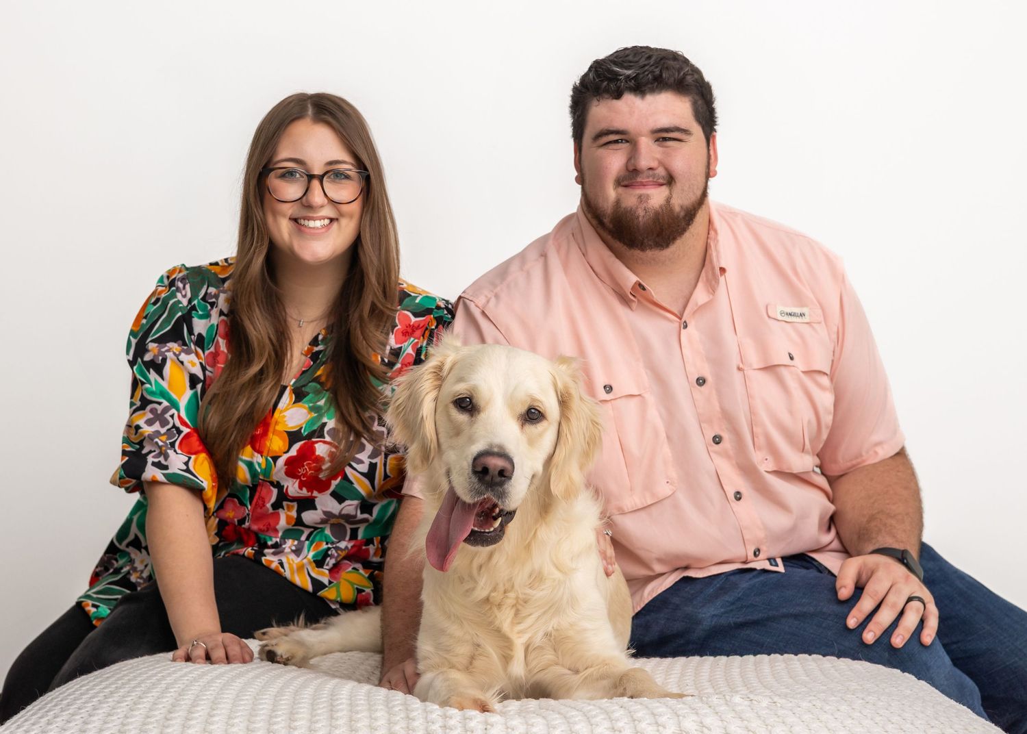 A golden retriever sits between two people on a white couch against a light background for a portrait session.