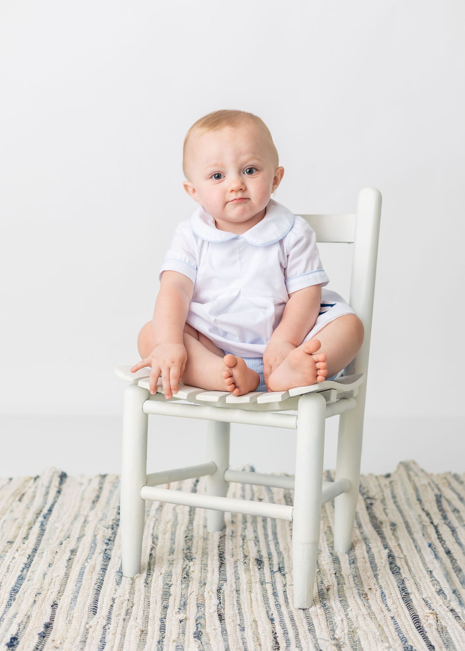 Baby in white outfit sitting on small white wooden chair against striped rug.