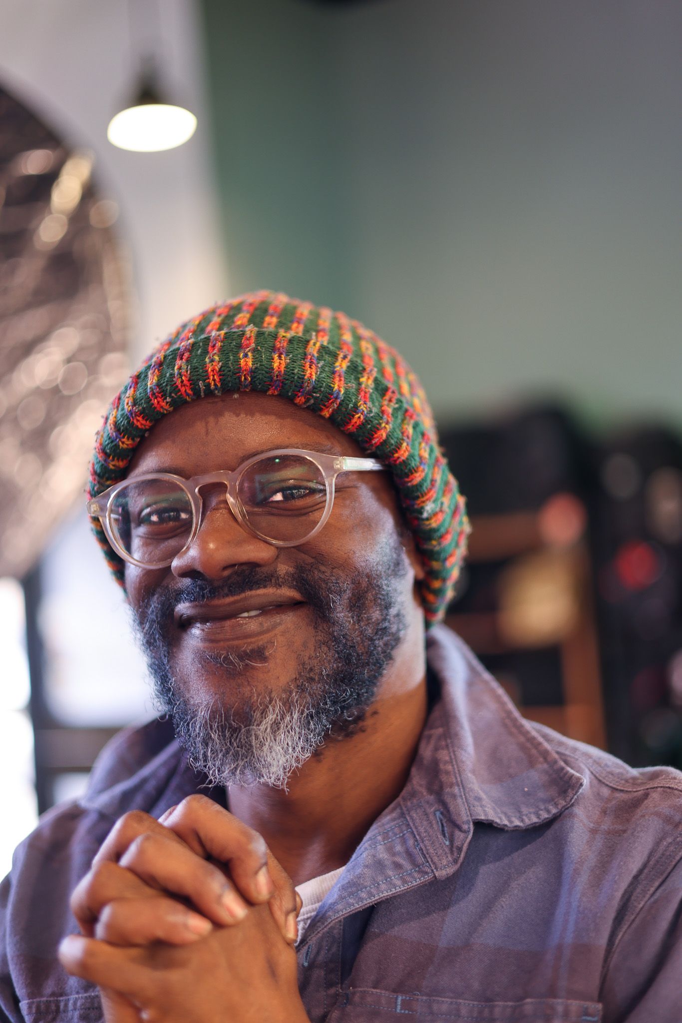 A person wearing glasses and a colorful knit beanie smiles warmly while clasping their hands together.