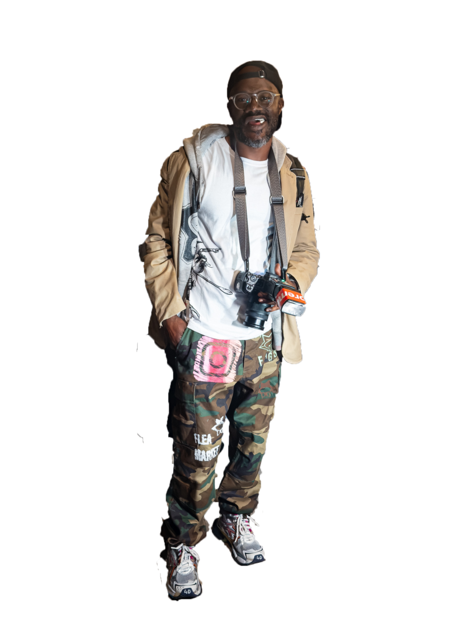 Man wearing camo pants, tan jacket, white shirt, and camera strap poses standing against white background.