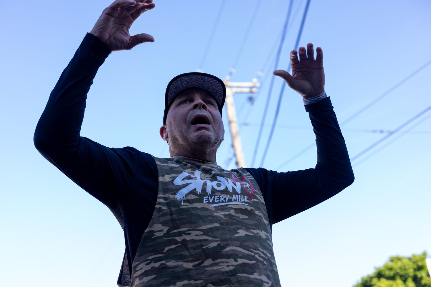 Man in camouflage shirt raises hands outdoors under clear blue sky.