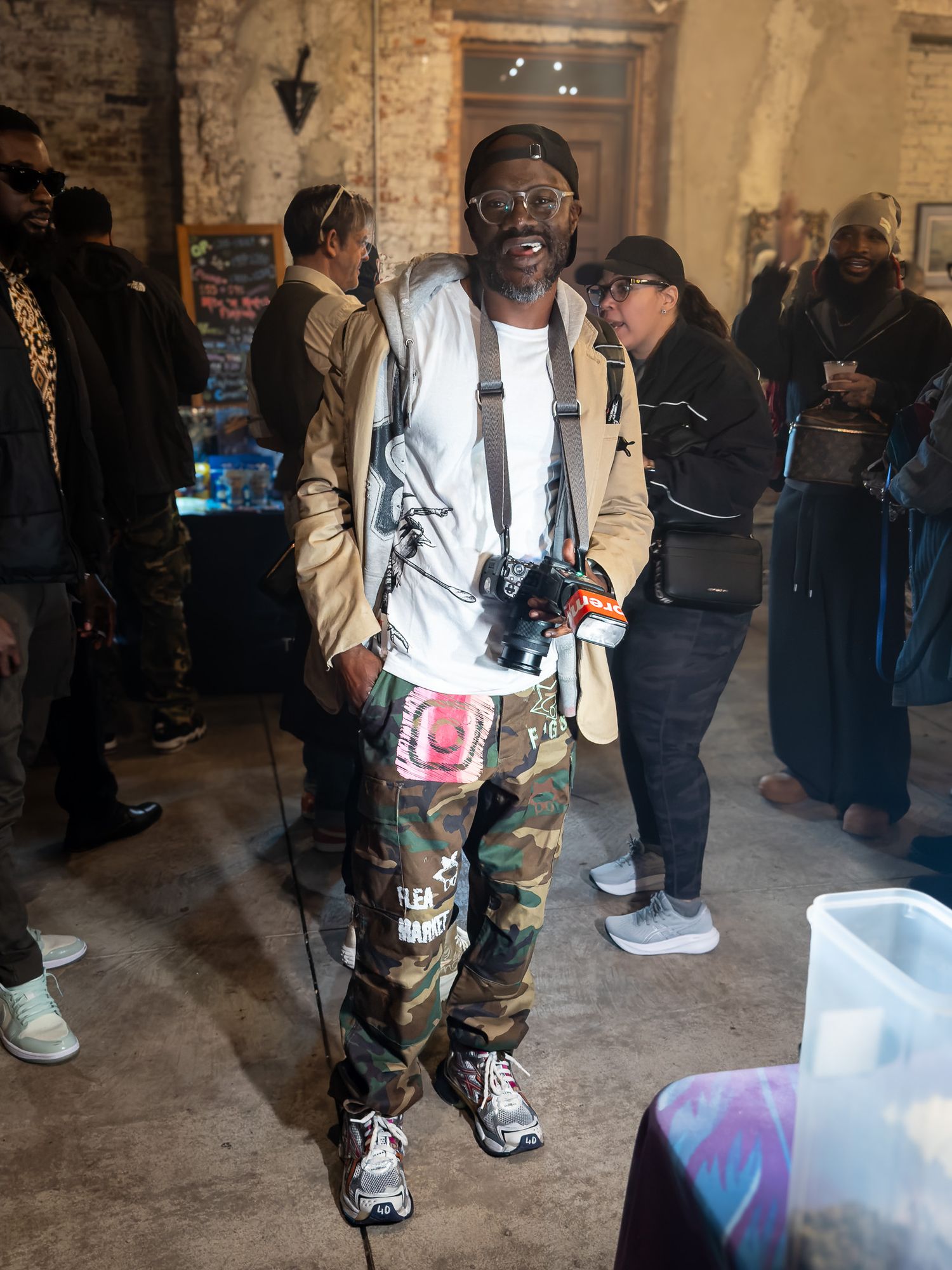 Labnek215 in person wearing camo pants and a light jacket stands in a brick hallway holding shopping bags.