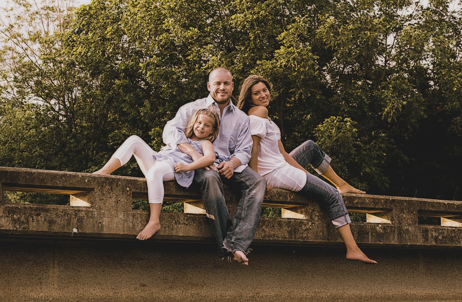 Family of three sitting together on a stone bridge surrounded by trees, barefoot, relaxed outdoor portrait session, Finger Lakes NY.