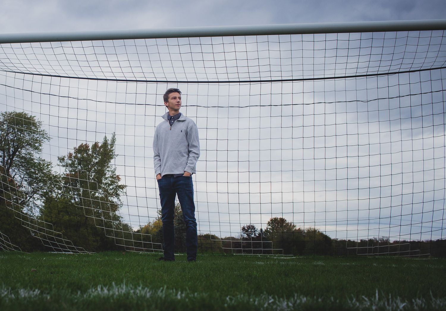 Teenager standing alone in a wide open field by a soccer goal, under a dramatic cloudy sky, outdoor portrait photography, Finger Lakes NY.