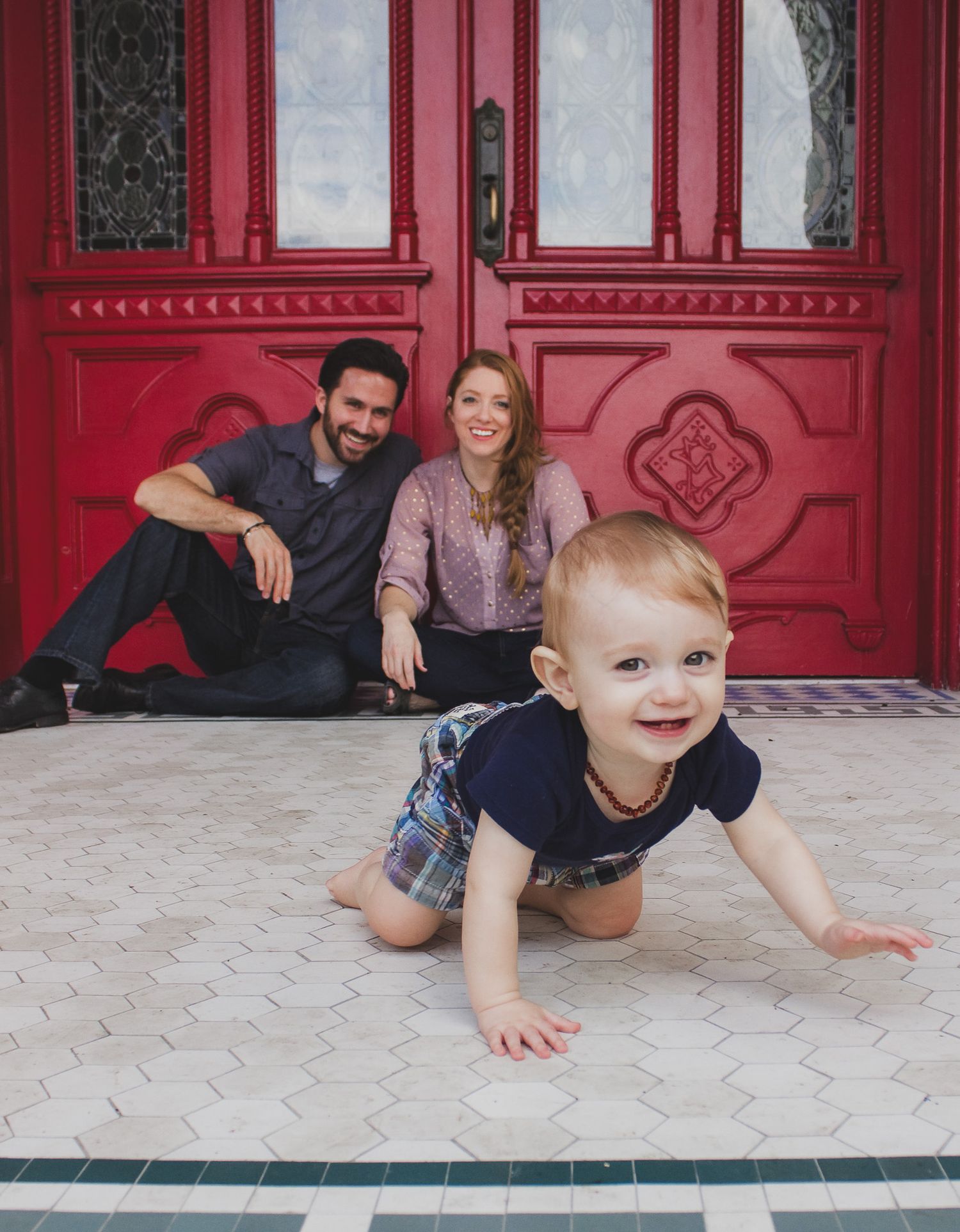 Family of three posed in front of a red door with an infant crawling towards the camera, natural light family portrait, Finger Lakes NY