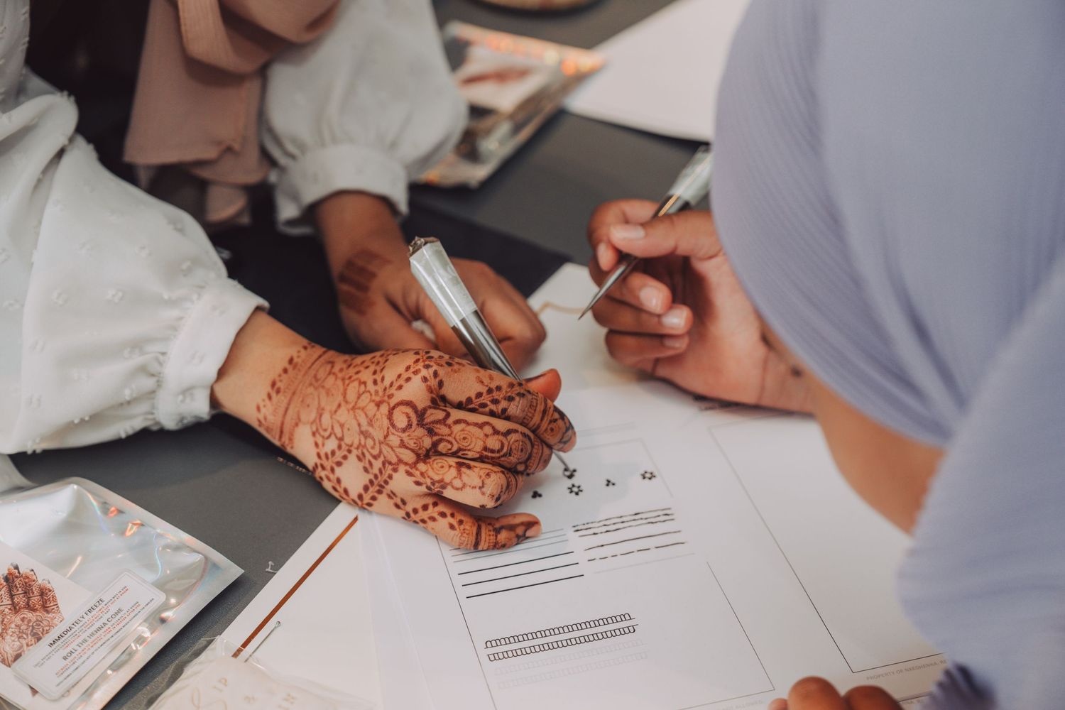 Close-up view of hands with traditional henna designs filling out paperwork at a desk.