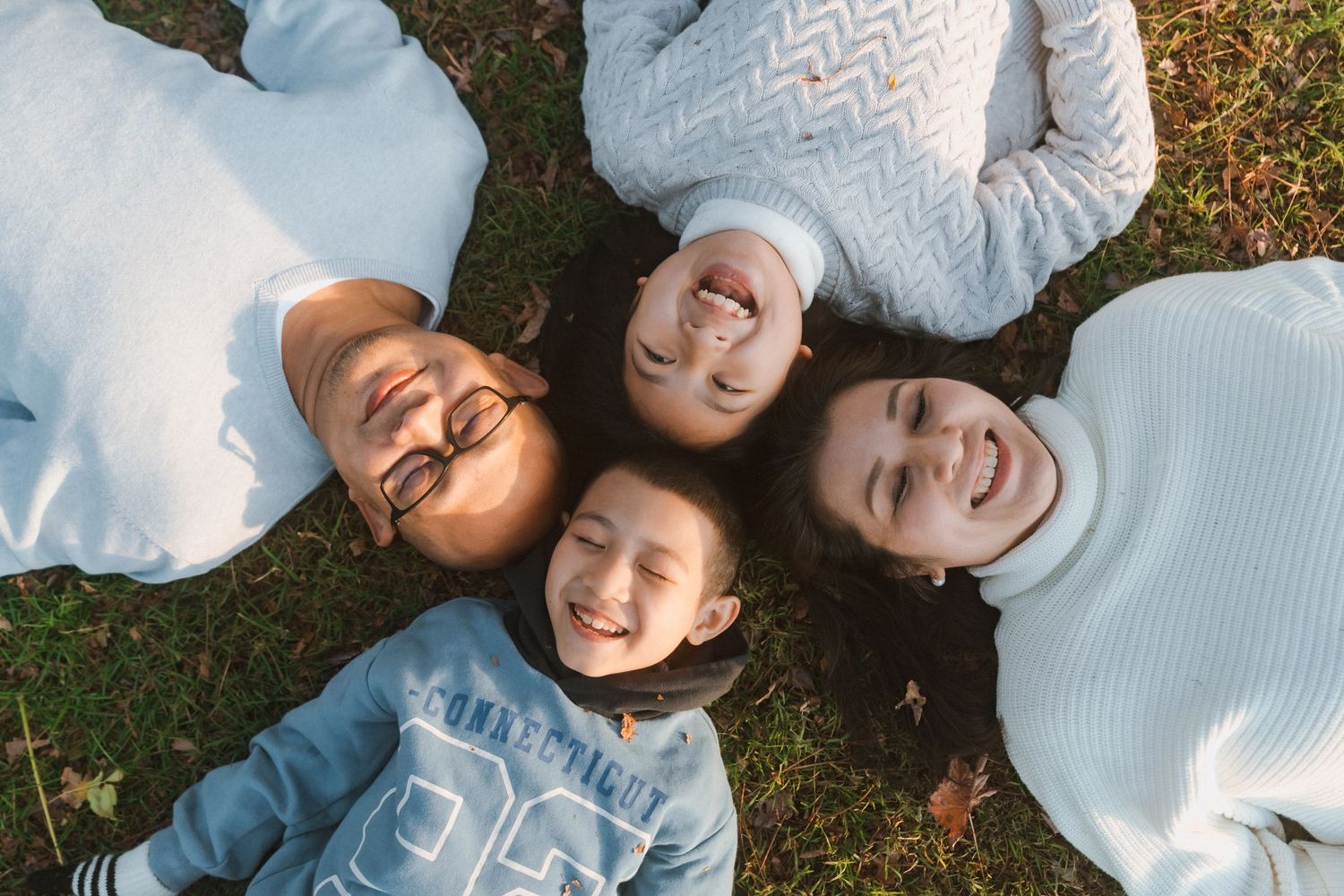 Overhead view of four people wearing light colored sweaters laying in a circle on grass with their heads together.