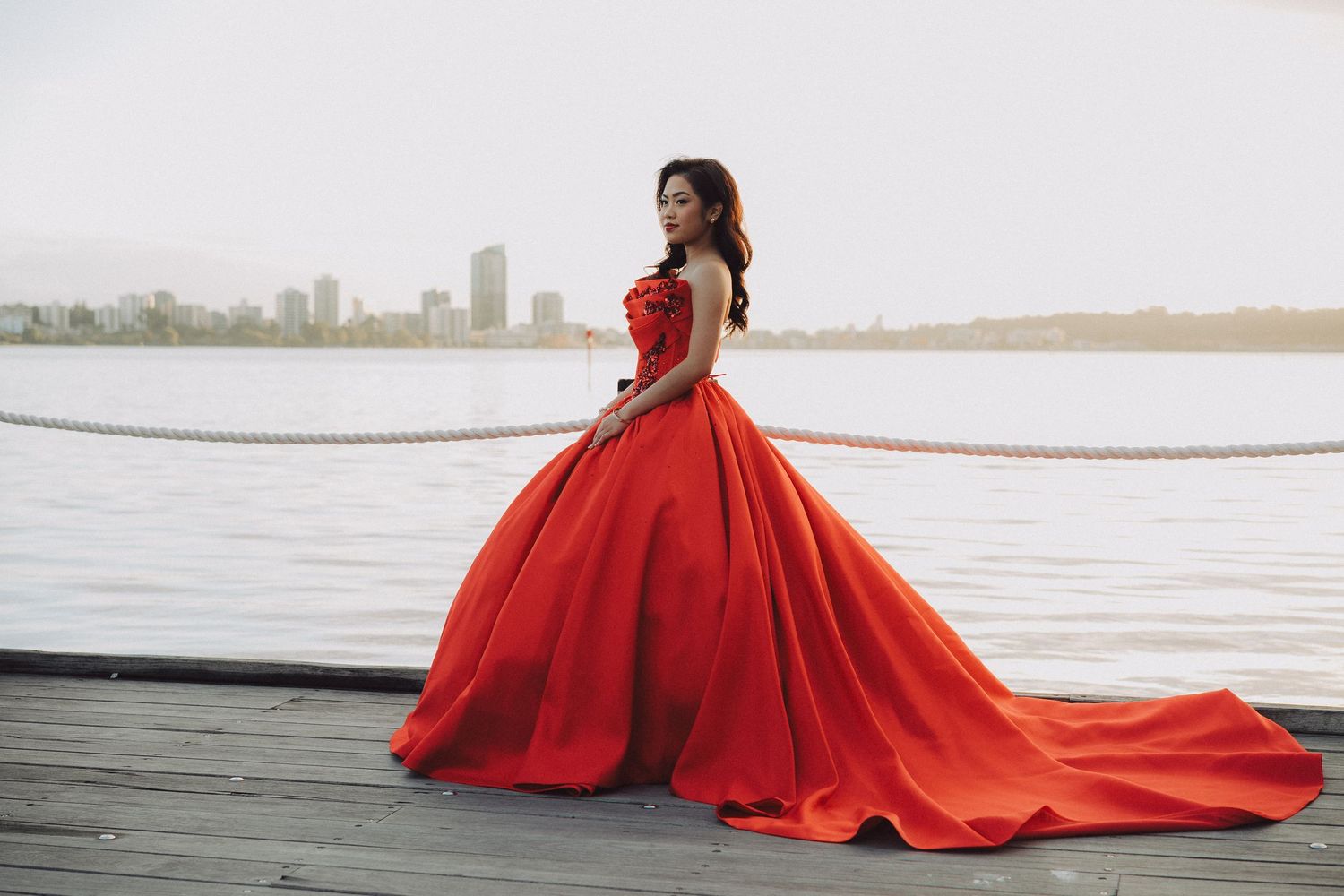 A model in a flowing red ball gown poses dramatically against a waterfront cityscape at sunset.