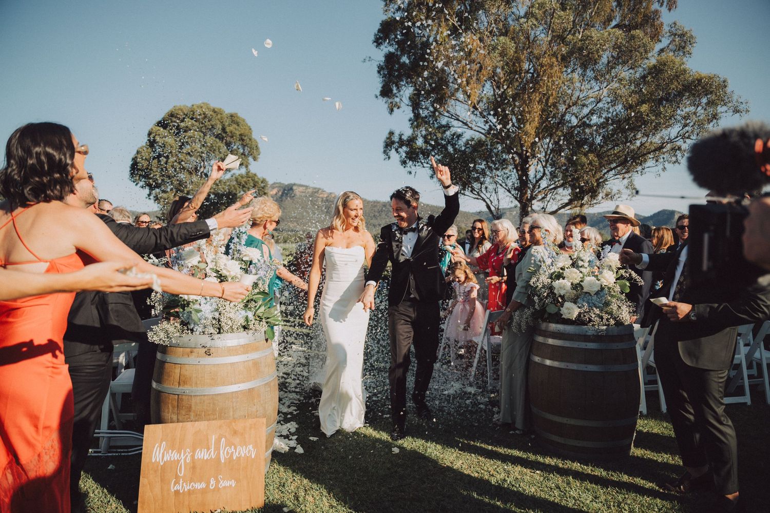 Wedding guests gather around wine barrels in a scenic vineyard setting during an outdoor celebration.