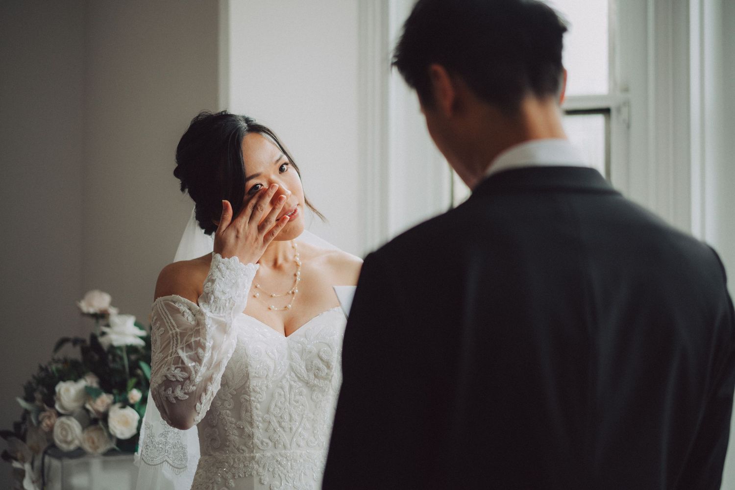 Bride in lace wedding dress covers her mouth in joyful emotion while standing with groom near window with flowers.