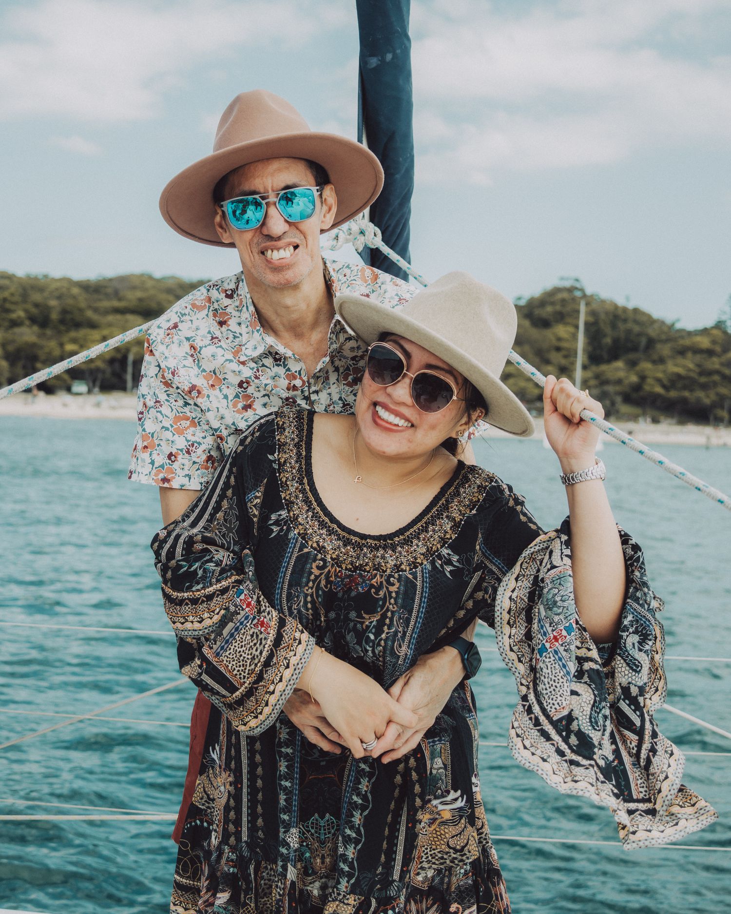 Woman in floral dress and sun hat poses joyfully on a boat with turquoise ocean waters and coastline in background.