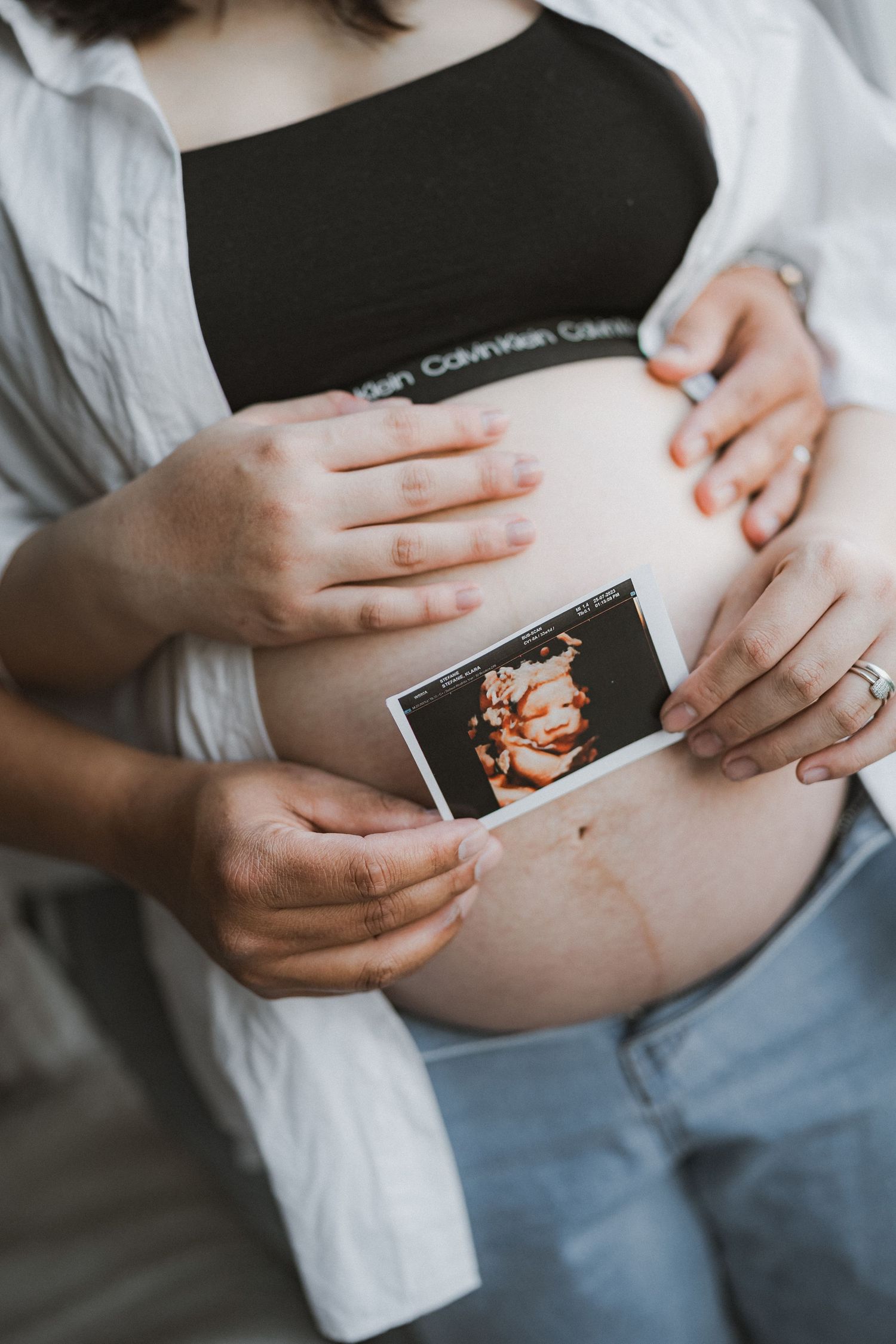 Hands hold a sonogram photo against a pregnant belly wearing a white shirt and blue jeans.