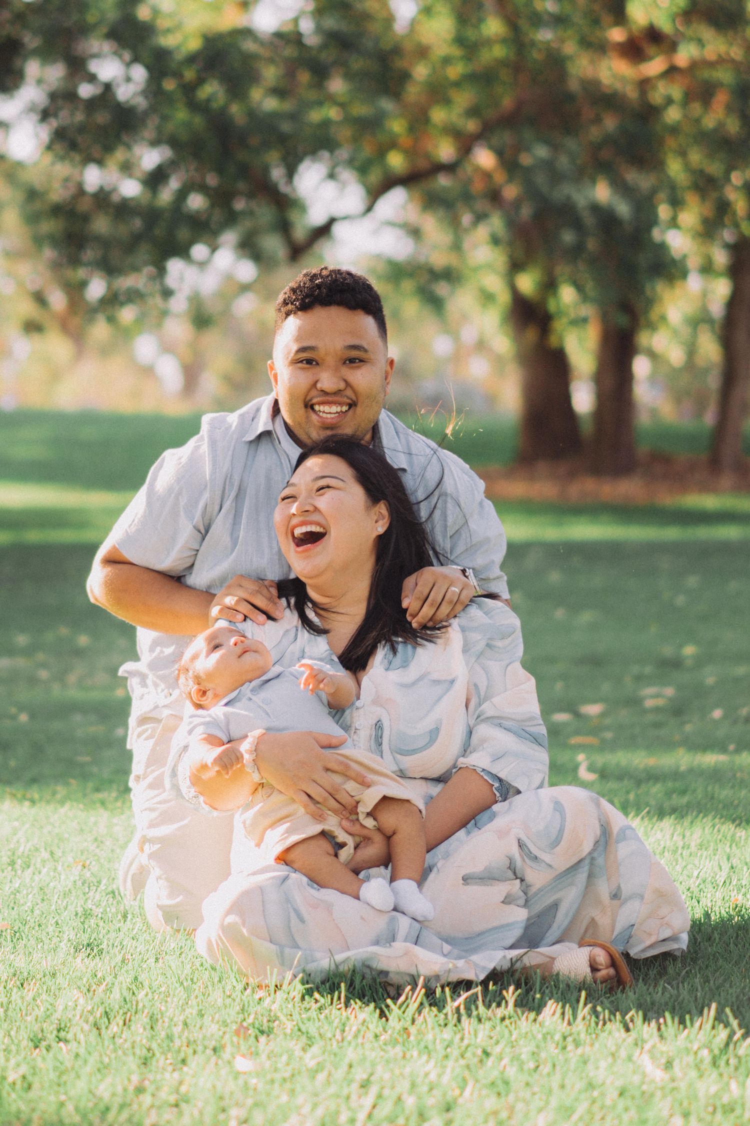 A couple in matching white outfits shares joyful laughter while relaxing together on green grass in a sunlit park.
