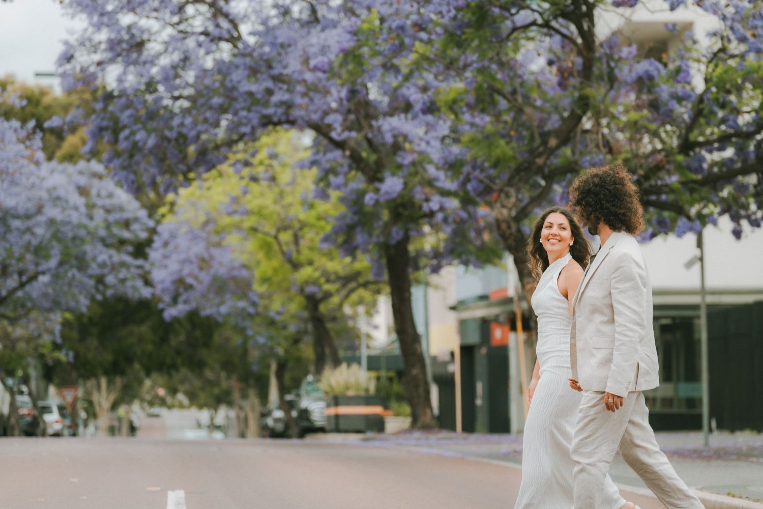A couple walks hand in hand beneath blooming purple jacaranda trees on a sunny spring day.