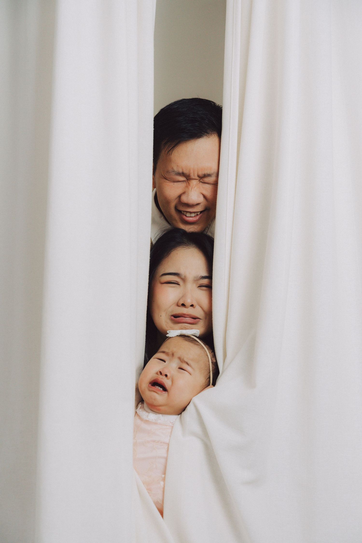 Three people peeking playfully through white curtains while making joyful expressions.