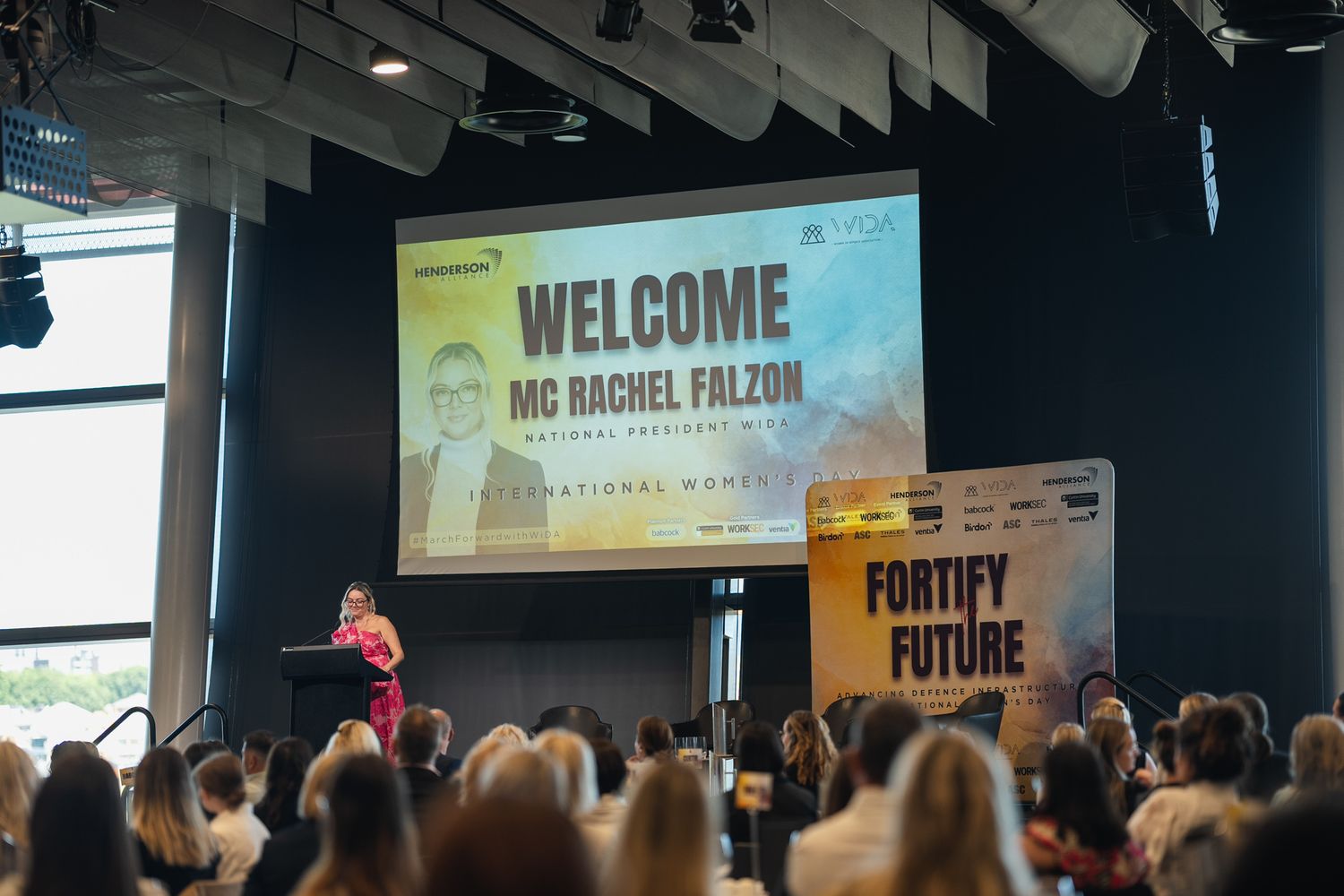 Conference attendees gather in a modern venue with welcome banners and presentation screens visible in the background.