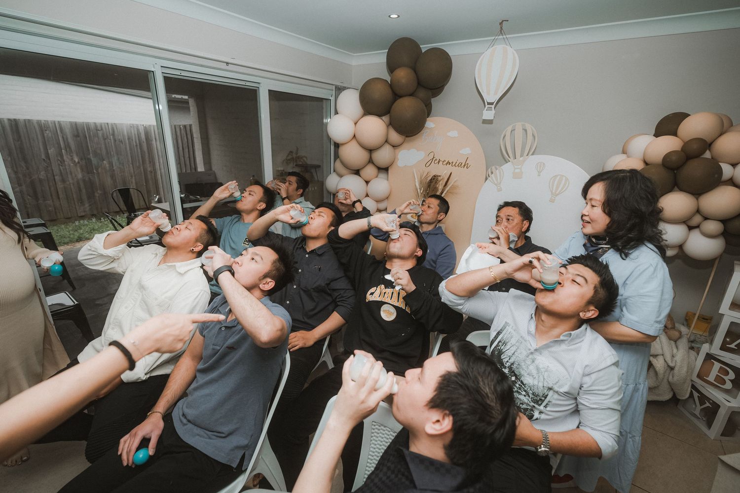 A group of party guests participate in a synchronized champagne toast at an indoor celebration with balloon decorations.