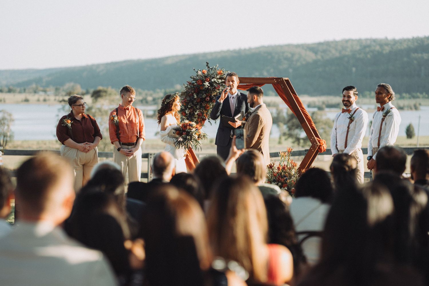 Outdoor wedding ceremony with couple at hexagonal arch, groomsmen, officiant, and lake mountain backdrop.