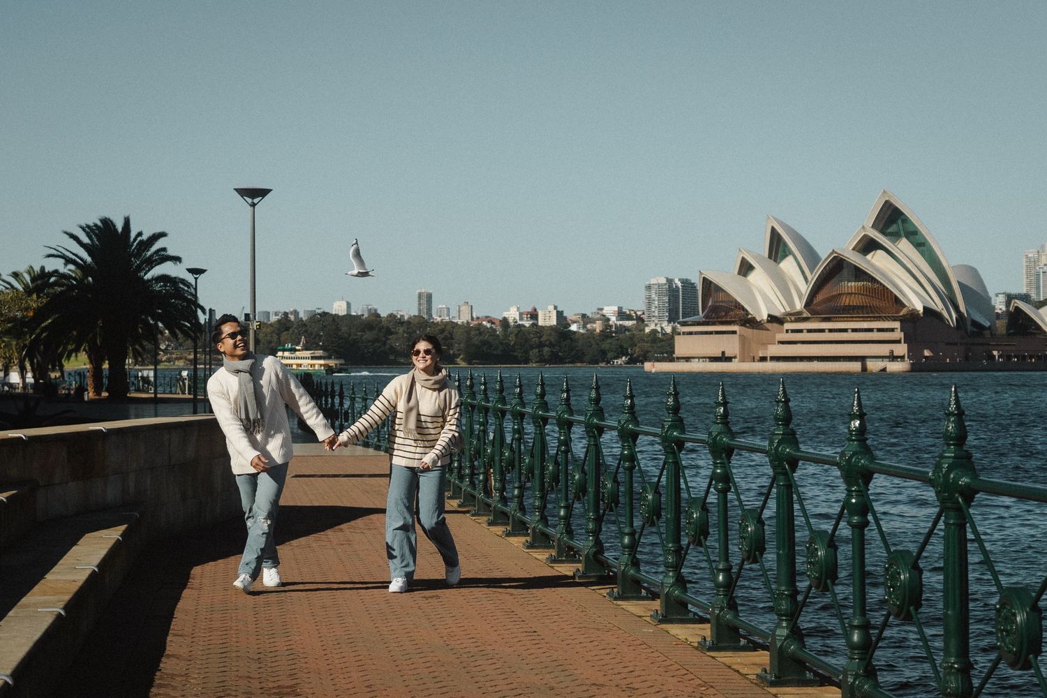 Two people walk along Sydney Harbour promenade with the iconic Opera House visible in the background.