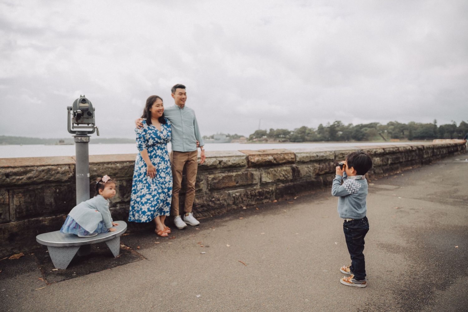 A family poses along a stone seawall on an overcast day at the beach while someone takes their photo.
