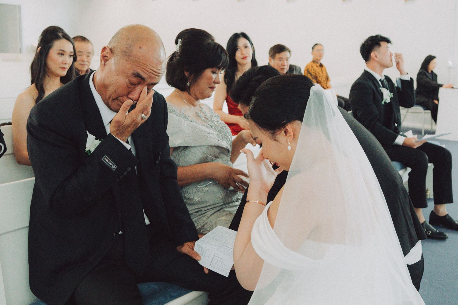 A wedding party sits together on a white bench during an emotional moment at a ceremony.
