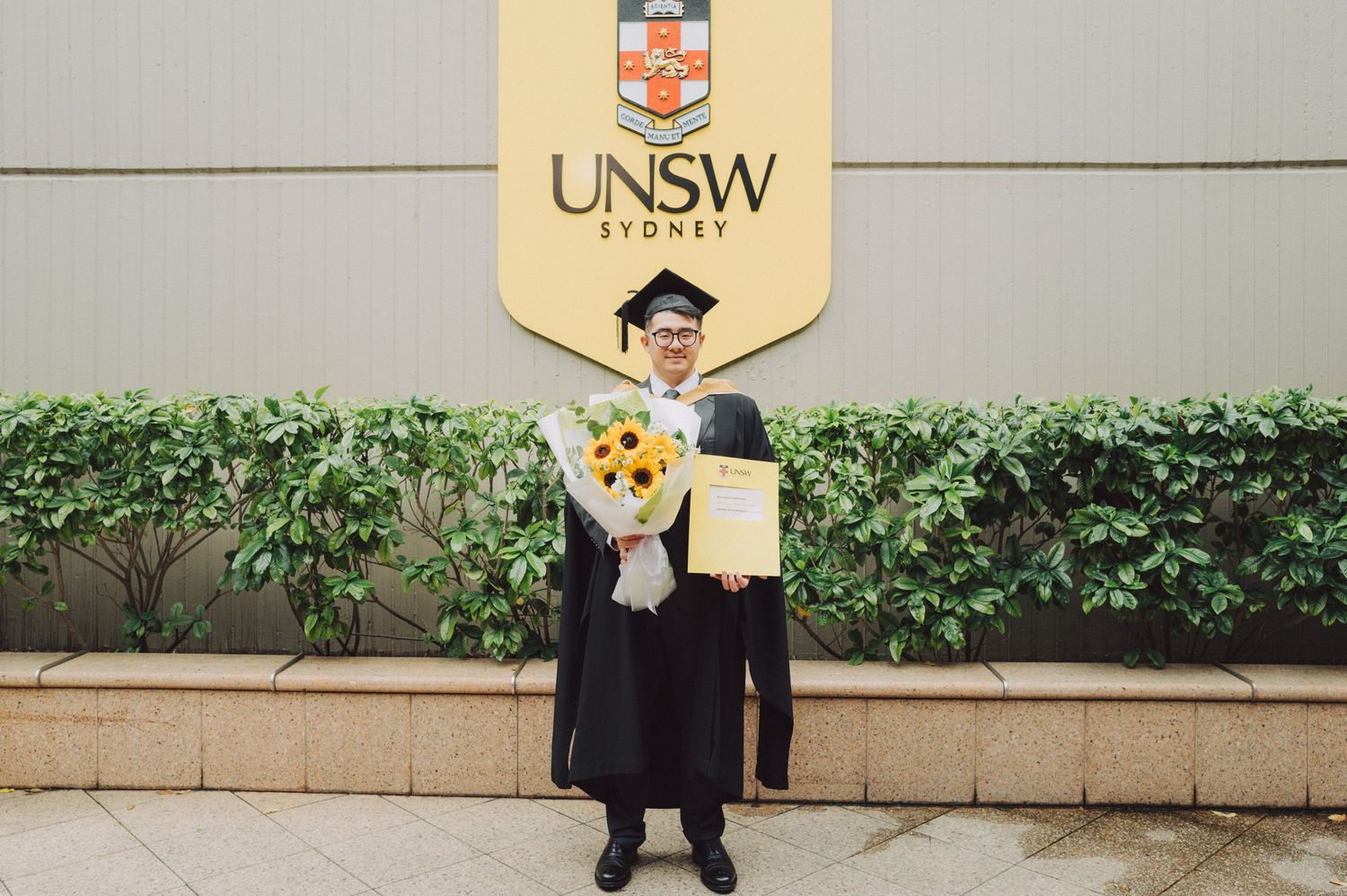Graduate in cap and gown poses for photos in front of a stone wall with greenery at UNSW.