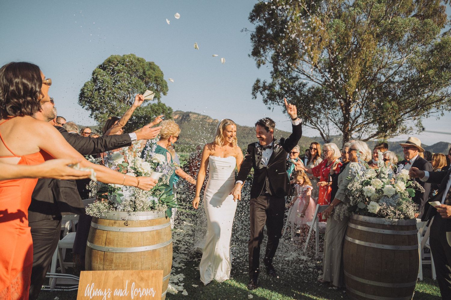 Guests throw confetti and celebrate at an outdoor winery wedding with wine barrels and trees in the background.